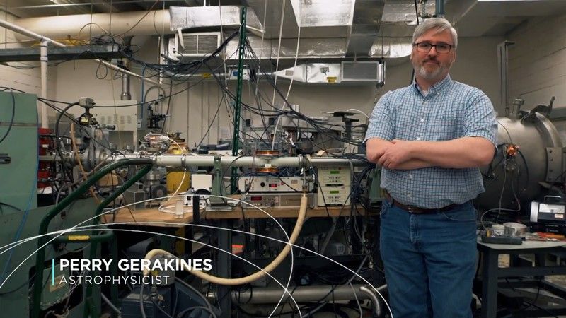 A man with White/gray hair and beard, posing in front of a piece of lab equipment