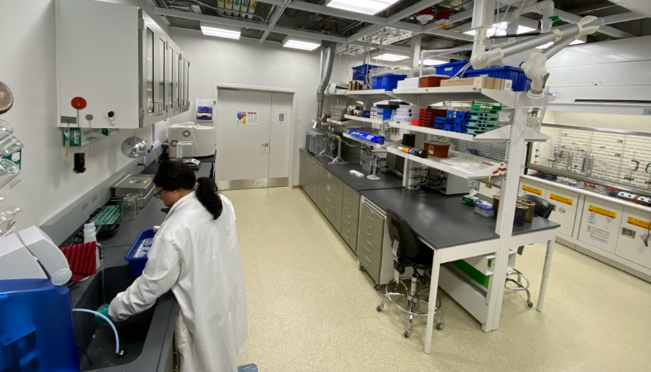 Woman in a white lab coat working in a laboratory. There are tables and shelves that reach the ceiling with various lab equipment on them.